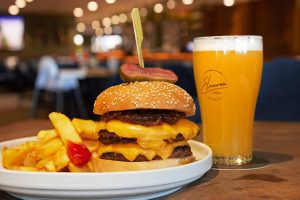 Picture is of chunky chips on a plate with a cheesy burger with a pickle on top, next to the burger is glass of beer with Elanora Hotel printed on the glass